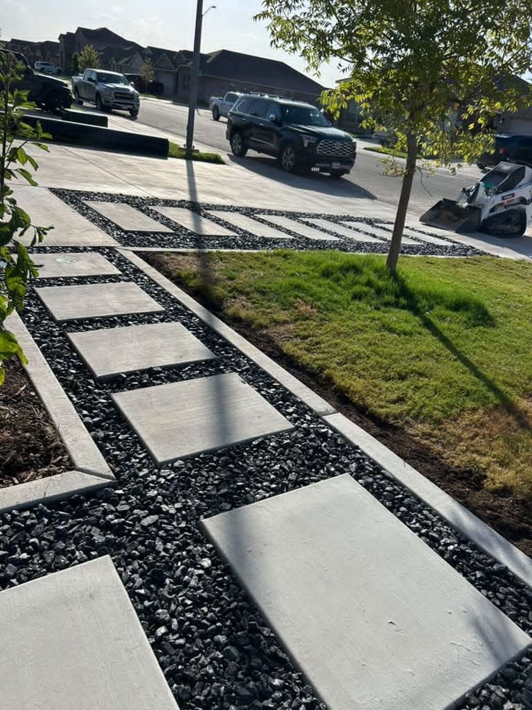Installation of a driveway and sidewalk using rectangular concrete slabs set on decorative black gravel, bordered by concrete edging and artificial turf in a residential neighborhood.