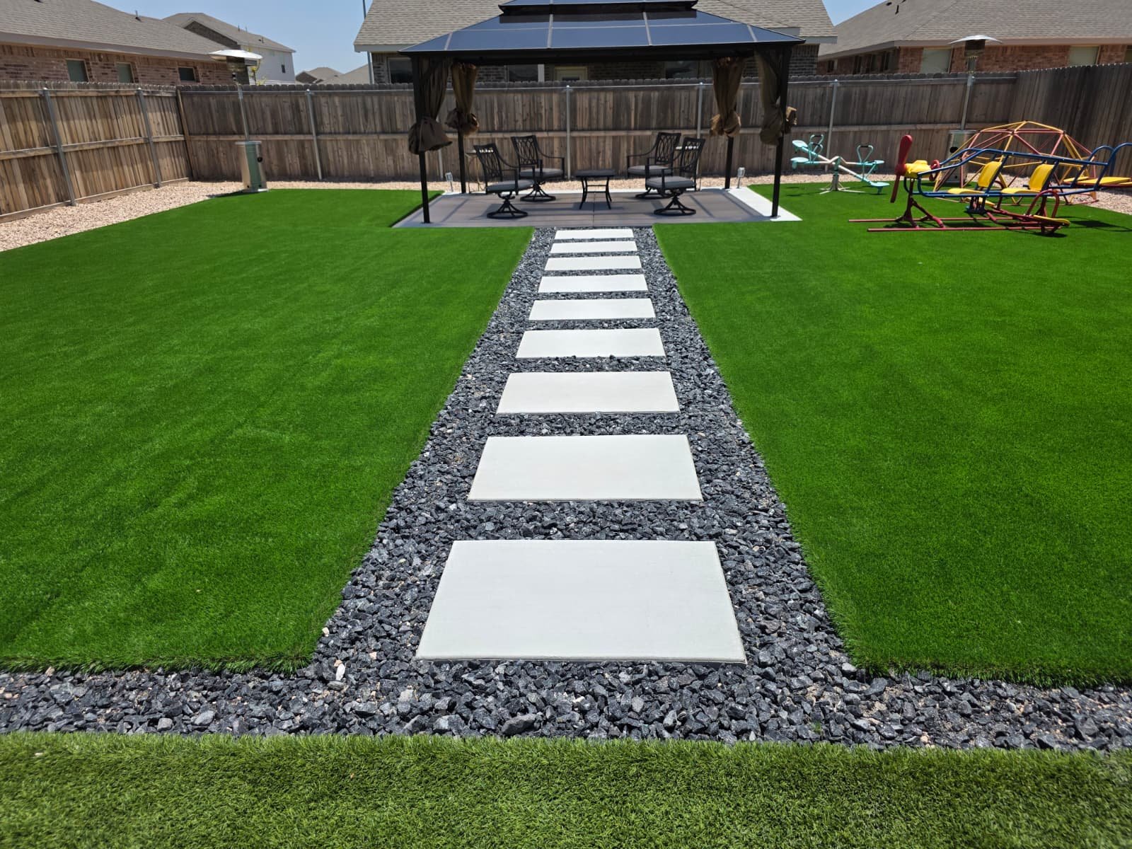 Residential backyard with professionally installed green artificial turf, a central path of white pavers flanked by dark gray gravel, a wooden pergola, and a children's play area.