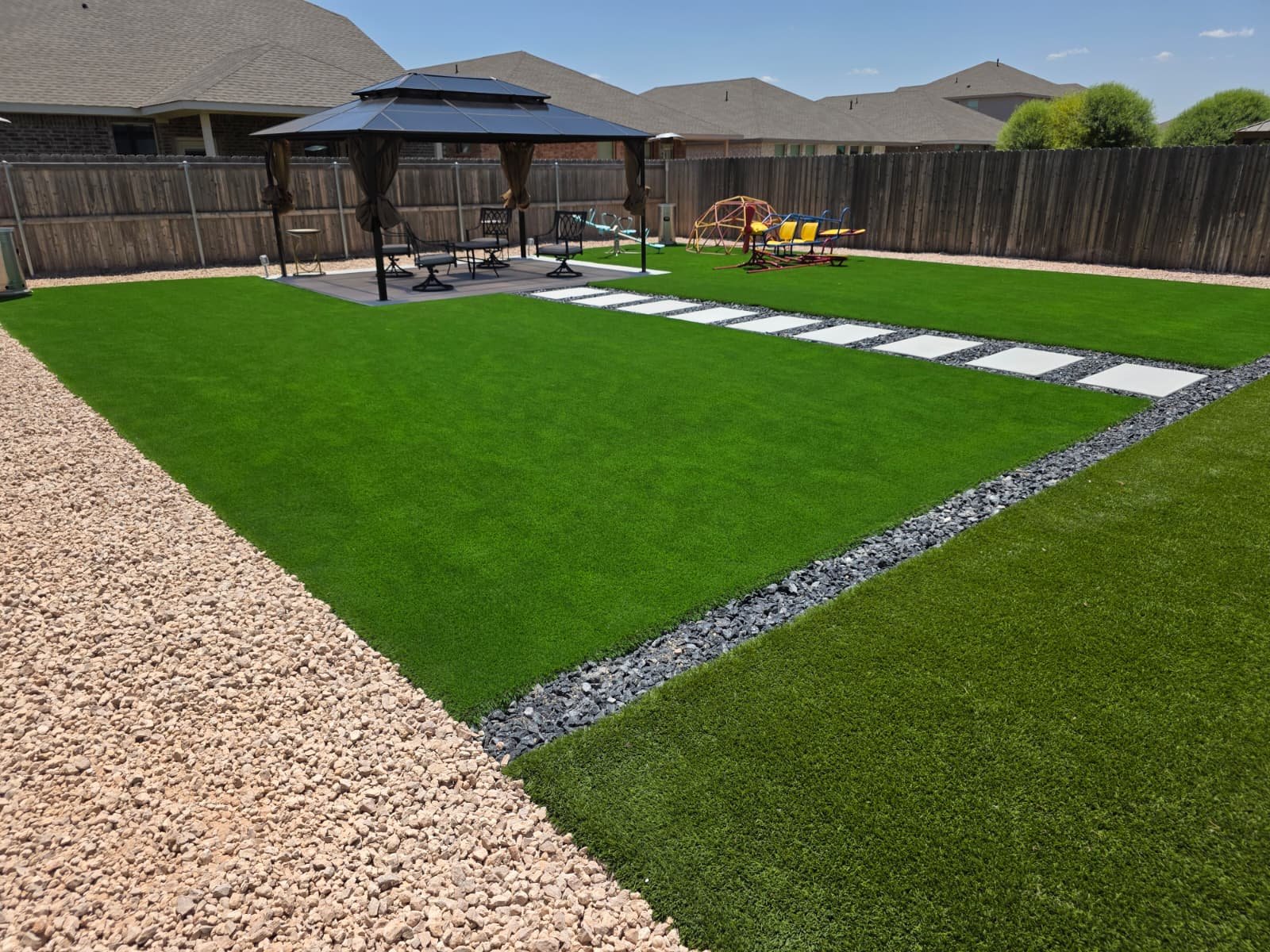 Professional installation of artificial turf in a residential backyard featuring a design with gray stone lines, a white flagstone path, and a decorative gravel border. Includes a pergola and a seating area.